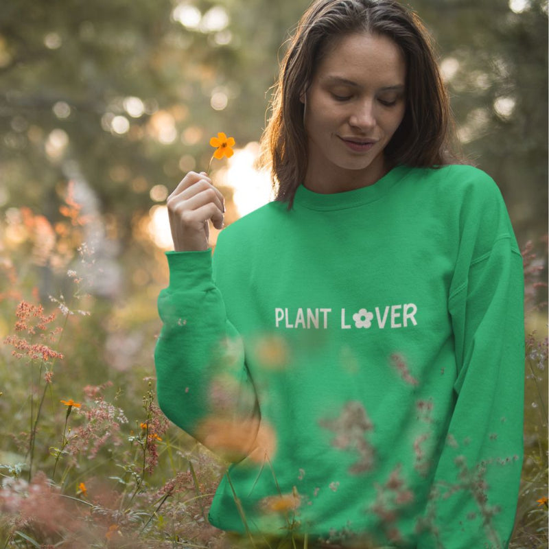 A woman wearing an irish green sweatshirt from Less Hustle, More Joy with the text 'PLANT LOVER' on the front in white, standing in a floral field.