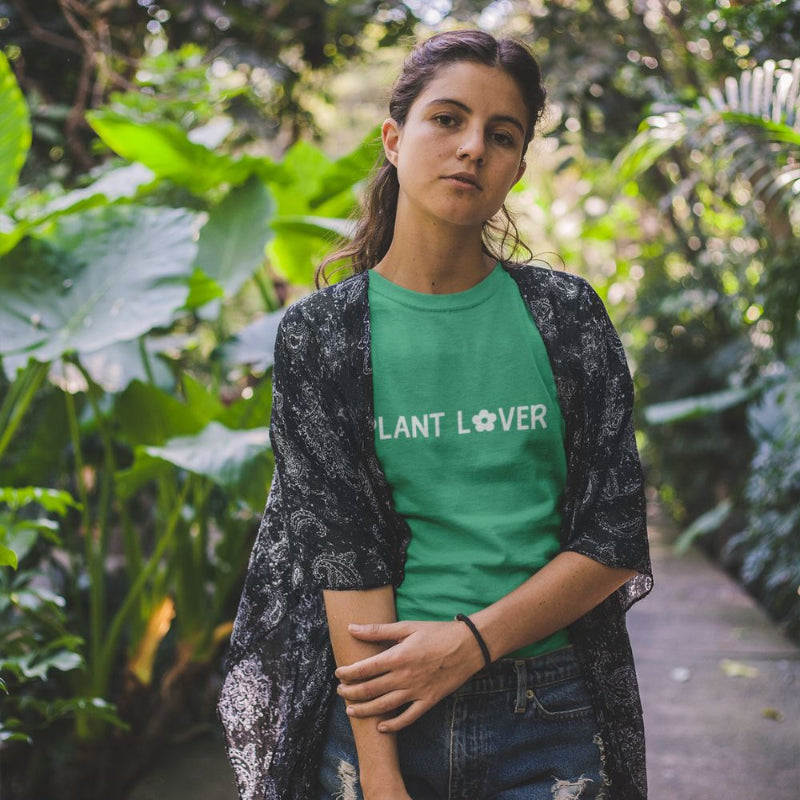 A person standing in a lush garden wearing a kelly green t-shirt by Less Hustle, More Joy, with the text 'PLANT LOVER' in white on it.