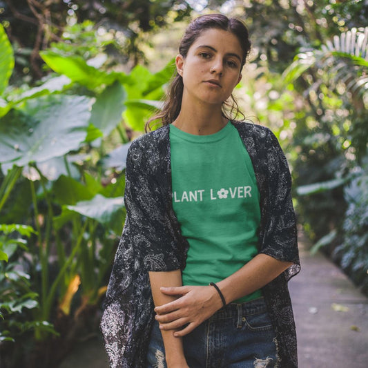 A person standing in a lush garden wearing a kelly green t-shirt by Less Hustle, More Joy, with the text 'PLANT LOVER' in white on it.