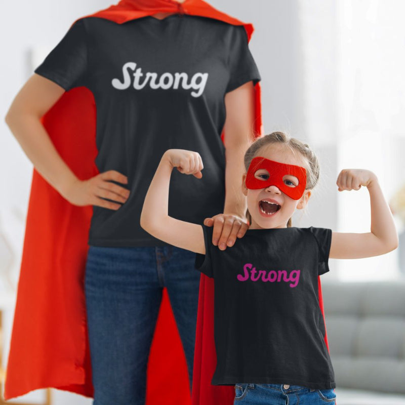 A toddler wears a superhero cape & mask with a black, short-sleeved toddler tee by Less Hustle, More Joy. The front features the word 'Strong' printed in punk rock pink text. In the background is an adult wearing a matching outfit.
