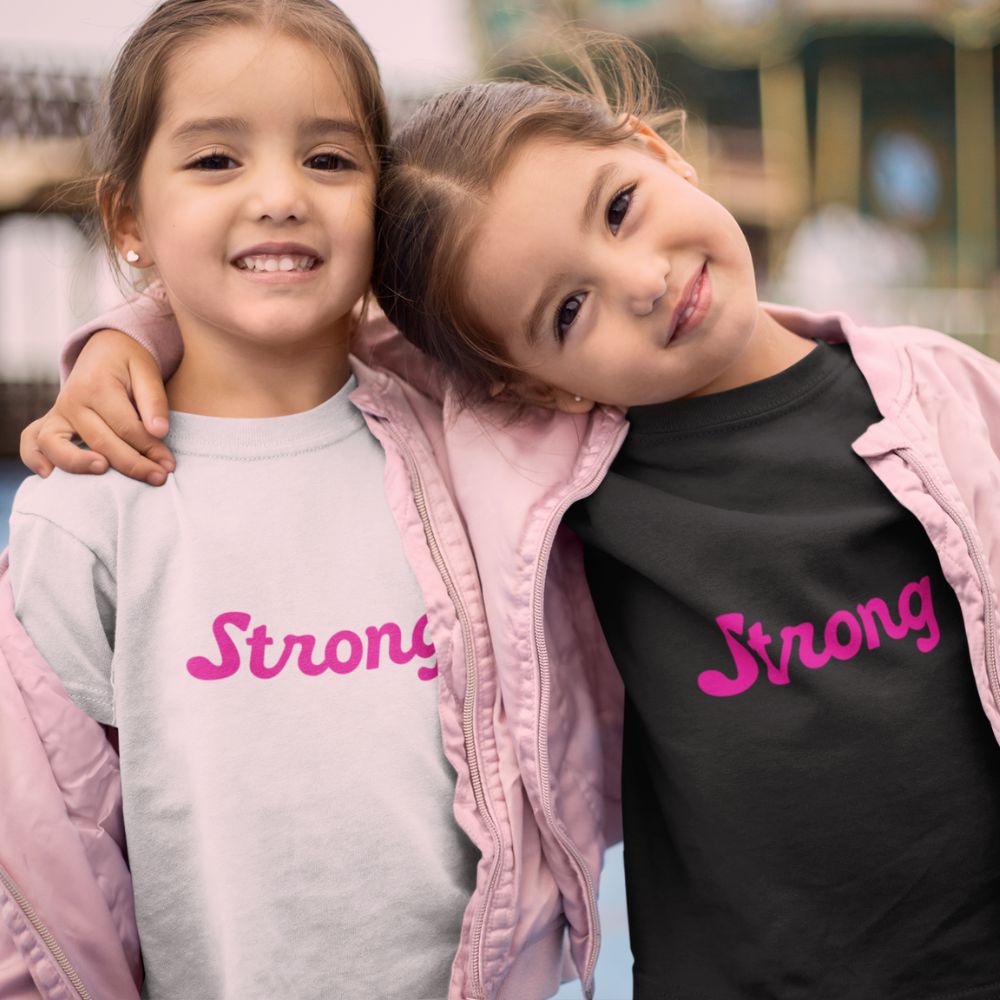 Two toddlers, one in a white t-shirt and the other in a black t-shirt with the word 'Strong' printed in punk rock pink, by Less Hustle, More Joy. Both toddlers are smiling and standing close to each other.