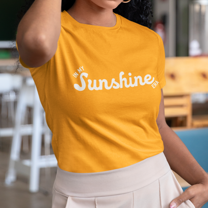 A person wearing a goldcrew neck t-shirt by Less Hustle, More Joy with the phrase 'In My Sunshine Era' printed in white text on the front.