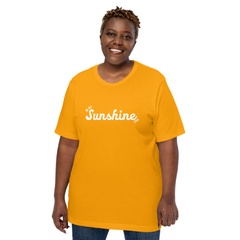 A person shown wearing a gold crew neck t-shirt by Less Hustle, More Joy with the phrase 'In My Sunshine Era' printed in white text on the front.