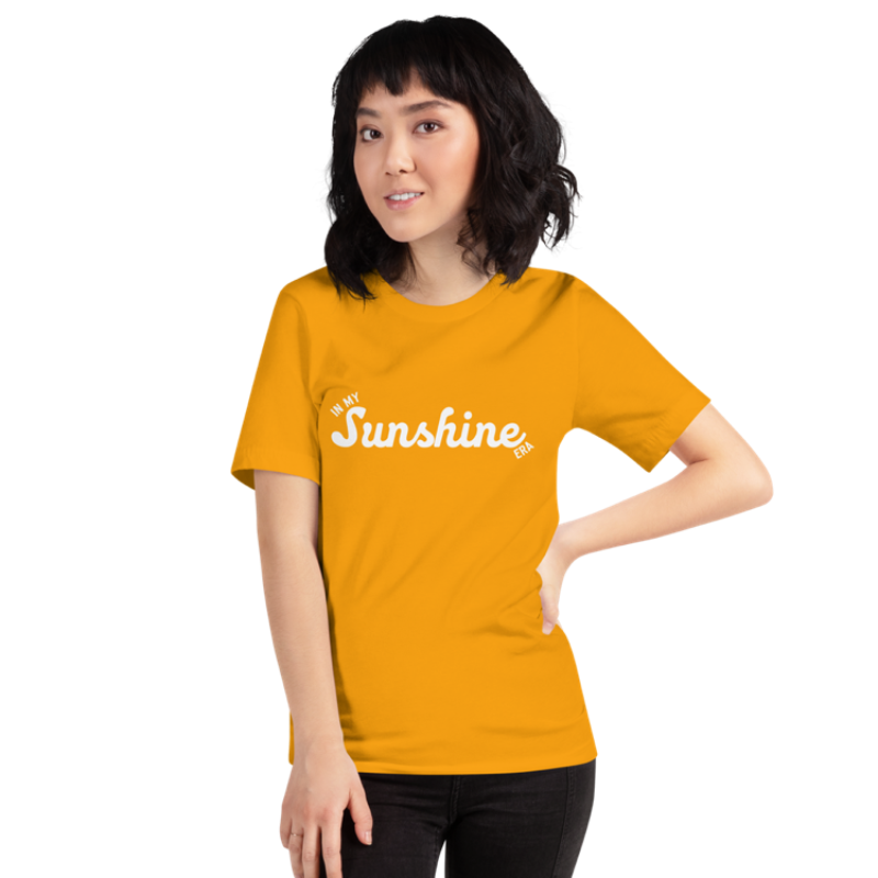 A person wearing a gold crew neck t-shirt by Less Hustle, More Joy with the phrase 'In My Sunshine Era' printed in white text on the front.