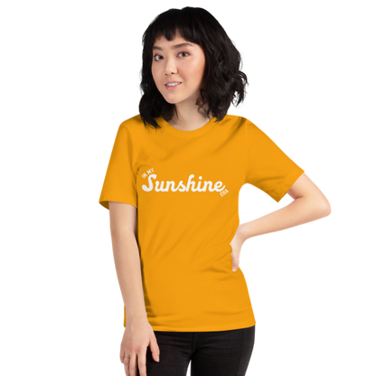 A person wearing a gold crew neck t-shirt by Less Hustle, More Joy with the phrase 'In My Sunshine Era' printed in white text on the front.