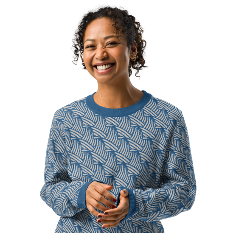A person smiling & wearing a crew-neck knitted sweater with a wave pattern in blue and sand colours by Less Hustle, More Joy.
