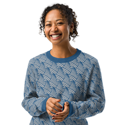 A person smiling & wearing a crew-neck knitted sweater with a wave pattern in blue and sand colours by Less Hustle, More Joy.